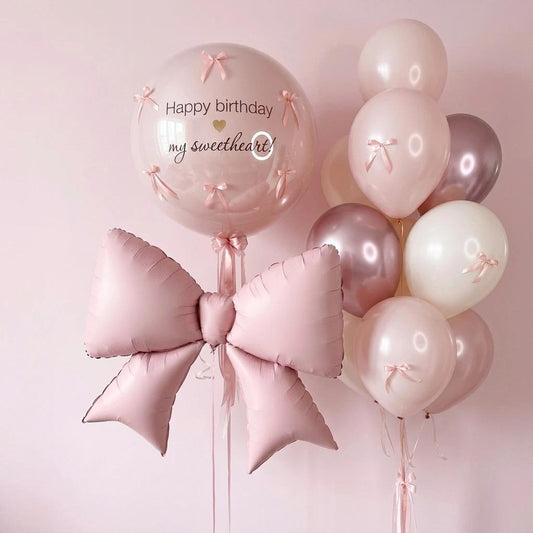 Pink birthday balloon bouquet with a large pink foil bow, a clear round balloon reading 'Happy birthday my sweetheart!' with small pink bow details, and a cluster of blush, cream, and rose-gold balloons against a soft pink background.